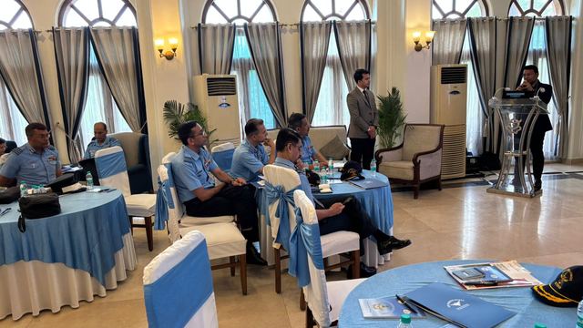 Officers in uniform seated at draped tables listening to a presentation, with caps set aside