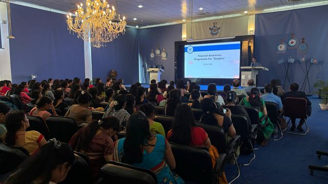Auditorium filled with attendees seated facing a presentation screen under a chandelier