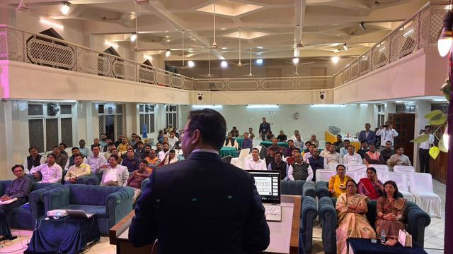 Speaker addressing a seated audience from a lectern in a double-height community hall